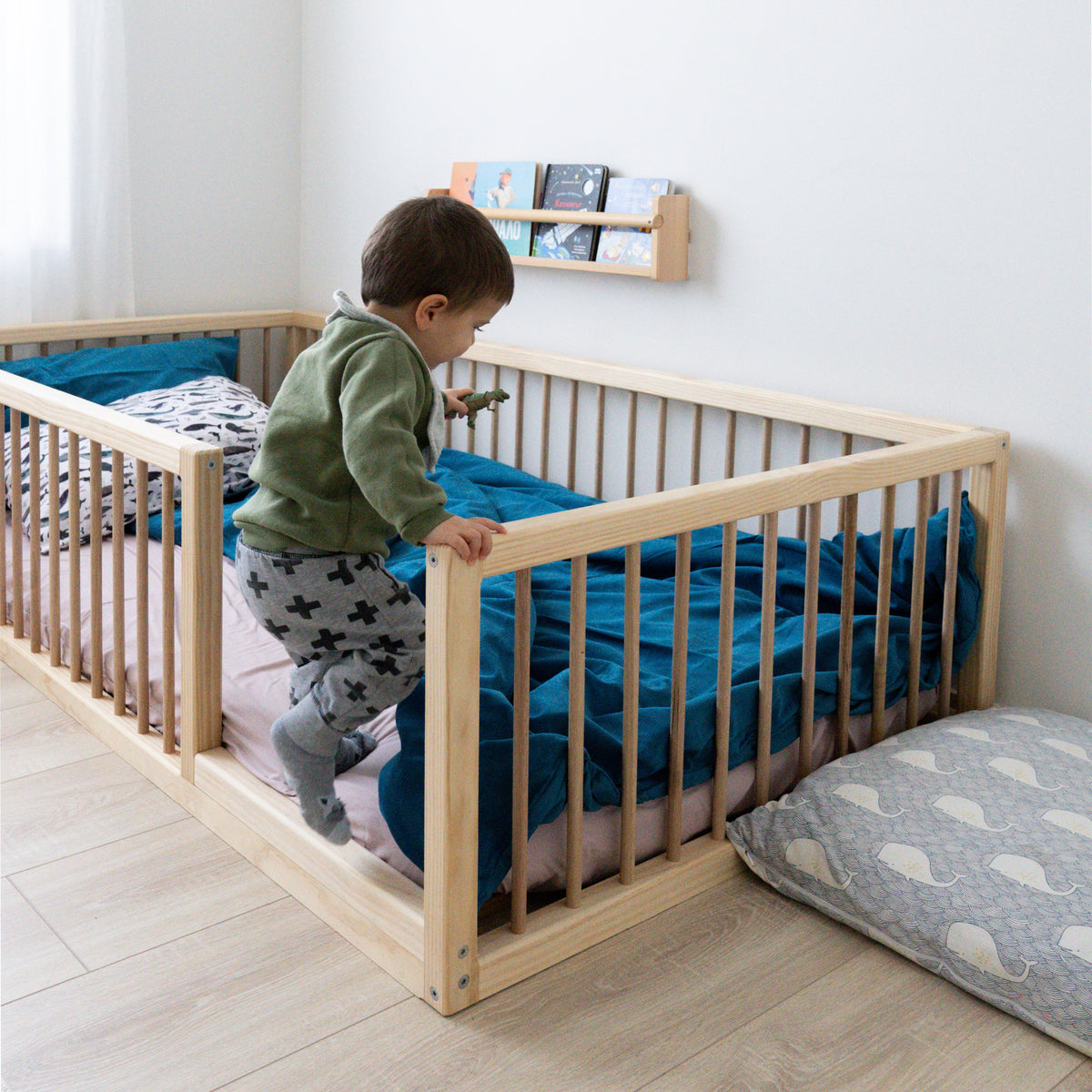 Child climbing into a wooden crib with blue bedding in a minimalistic room.